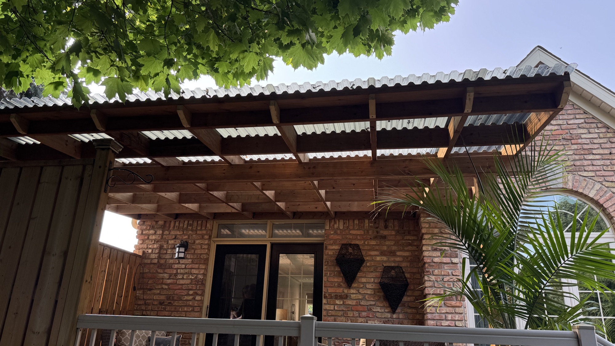 Deck roof framing seen from below against a brick Niagara home, showing cedar beams and clear polycarbonate panels