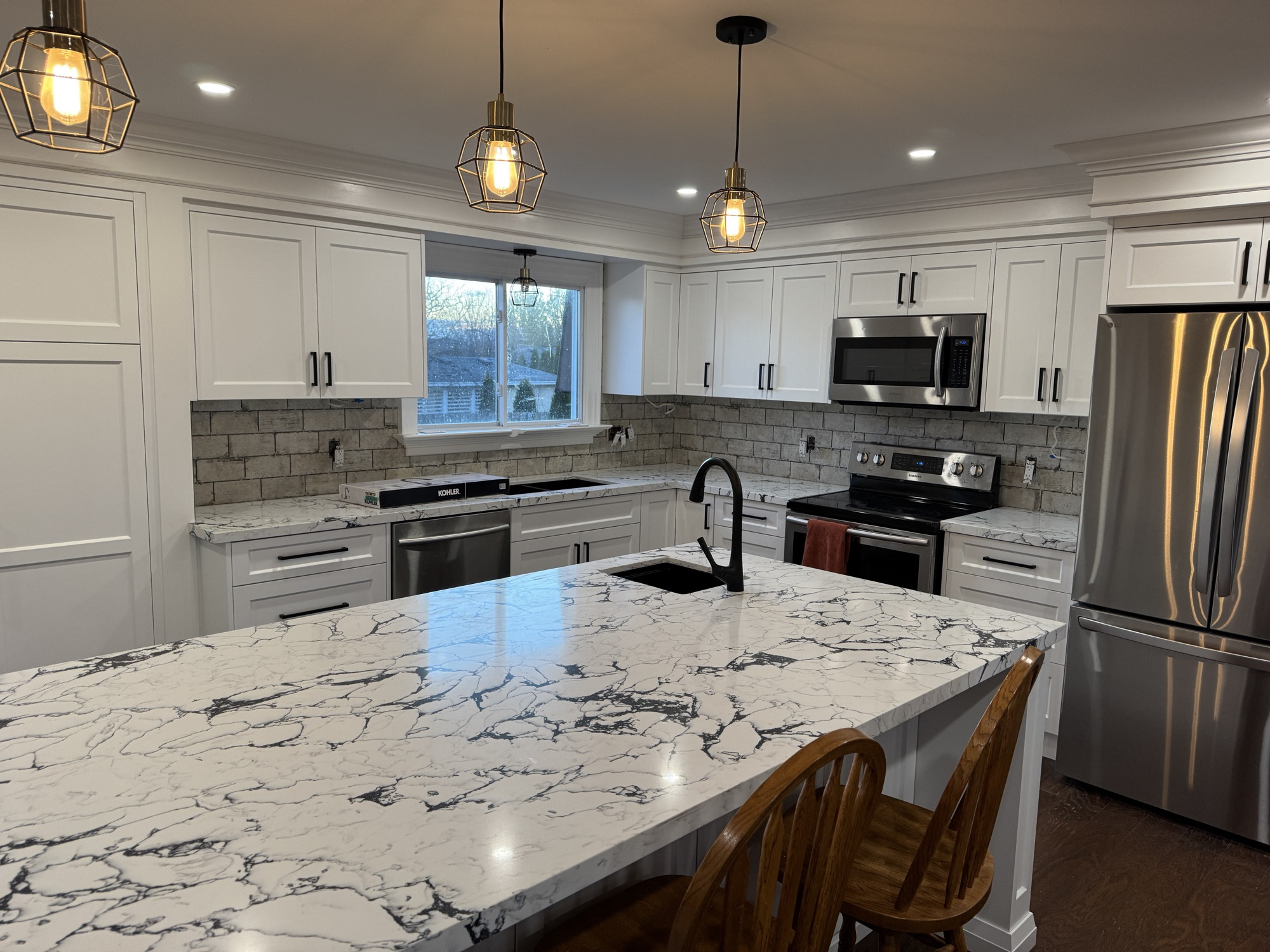 Renovated white kitchen with brass cage pendant lighting over a large marble island and stone subway tile backsplash